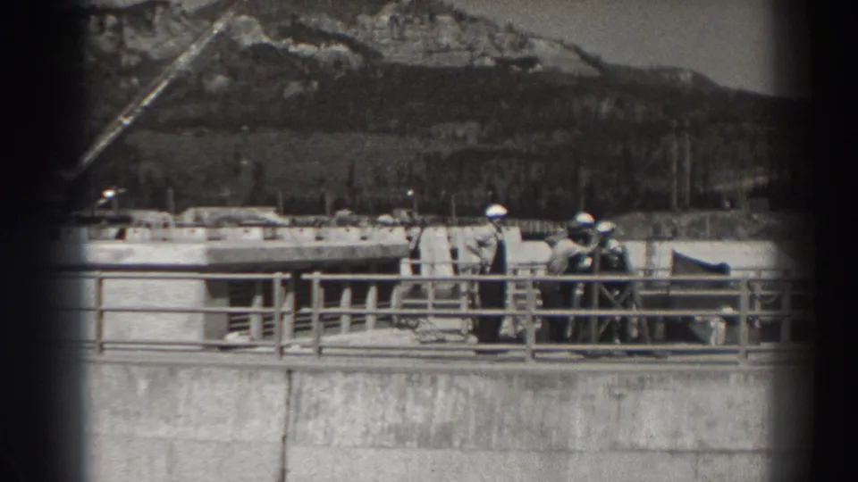 At Bonneville Dam in Oregon, several men in hard hats stand on a concrete structure with railings, industrial forms, and hills in the background.