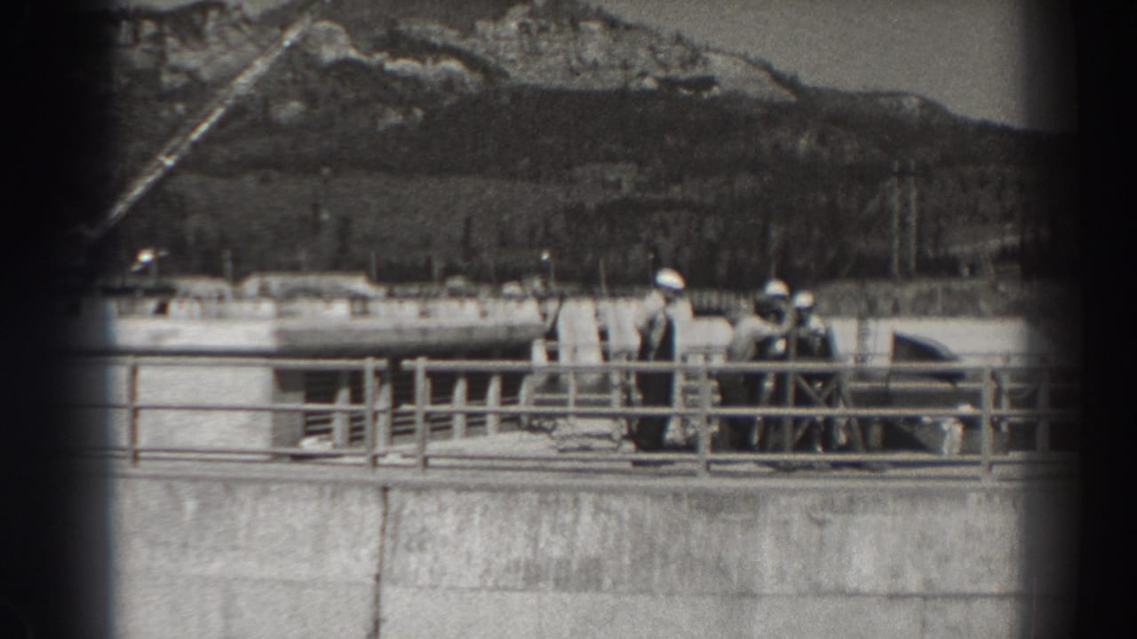 A small group stands near a railing on a concrete dam structure with construction equipment and hills beyond.