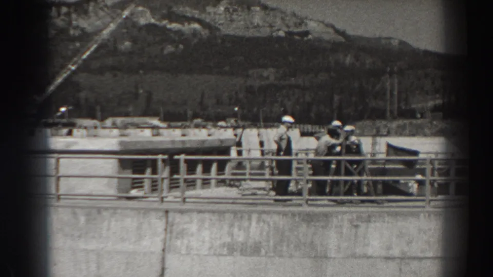 A grainy 8mm frame from Bonneville Dam shows men in hard hats standing by a concrete railing with construction features and hills in view.