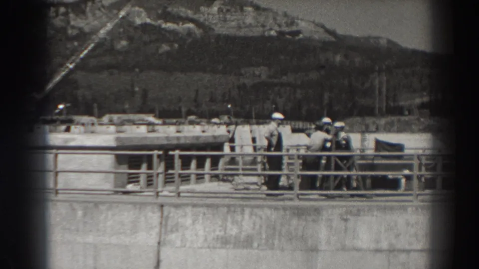 Bonneville Dam footage shows men standing together on a concrete structure, with a railing, small flag, dam works, and wooded hills behind them.