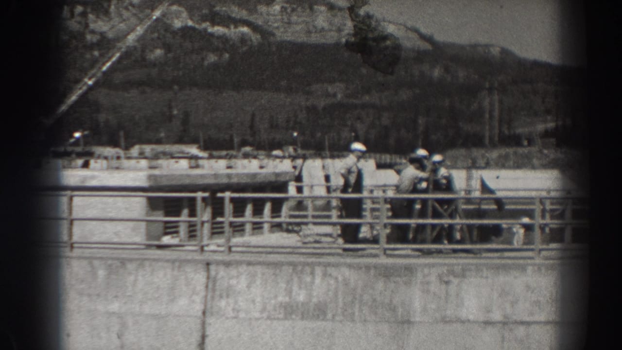 Hard-hatted men stand by railings on a concrete dam structure with hills behind them.