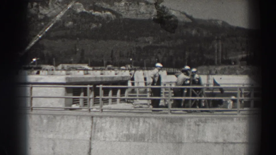 1938 Bonneville Dam view showing hard-hatted men on a concrete structure with railings and a mountainous backdrop.