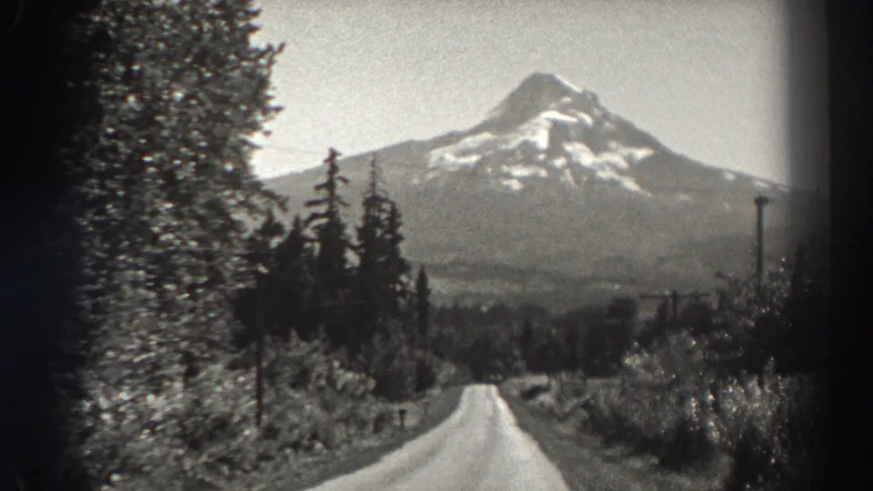 Oregon mountain-road scenery from the 1938 clip, with trees, utility poles, and a snow-capped peak beyond the road.
