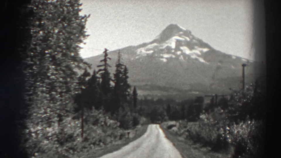Black-and-white 8mm view of a road descending through forest toward a snow-capped mountain in Oregon scenery.