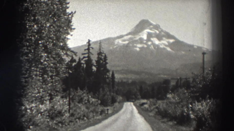 Late-1930s Oregon travel scenery with a road, dense trees, utility poles, and a snow-capped mountain peak.