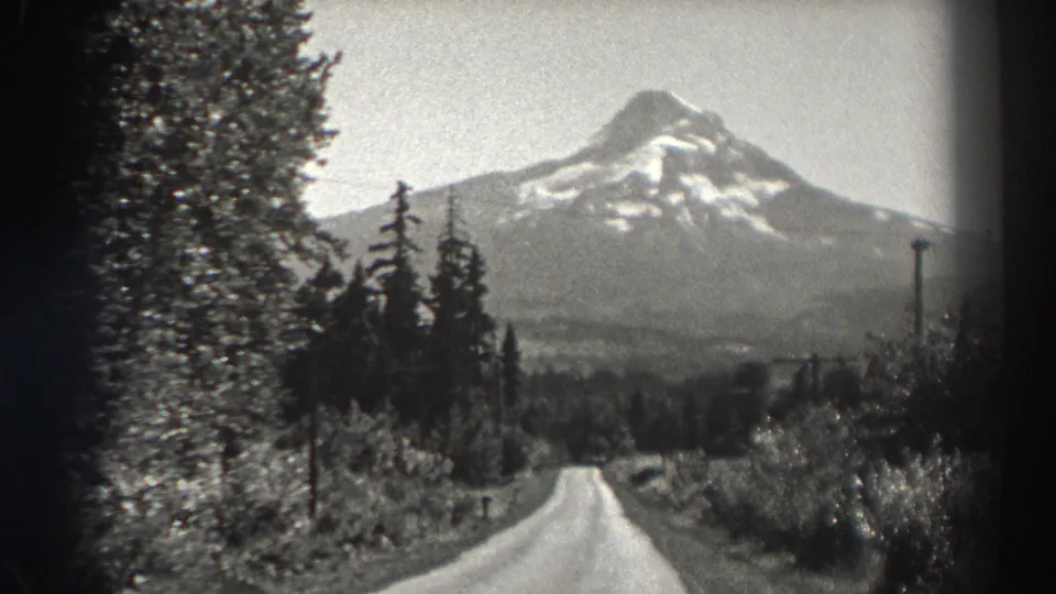 1938 Oregon home-movie frame showing a snow-capped mountain beyond a road lined with evergreens, brush, and utility poles.