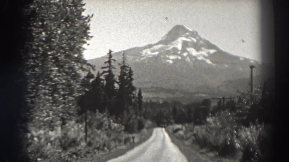 1938 Oregon frame with a snow-covered mountain rising beyond a tree-lined road and utility poles in a rural landscape.