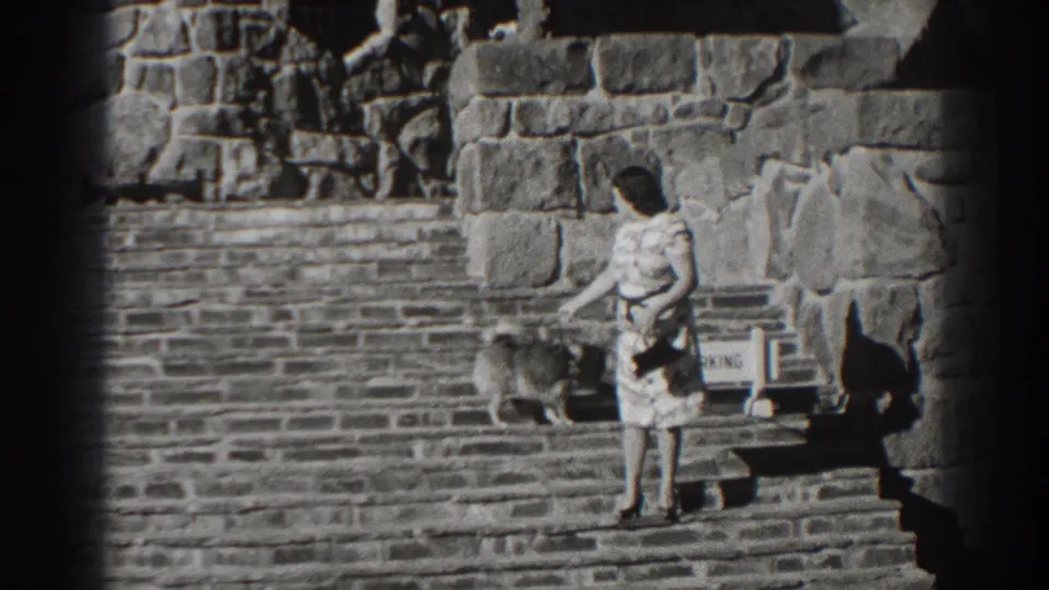 1938 Oregon home-movie frame of a woman interacting with a small dog on stone steps near Bonneville Dam masonry.