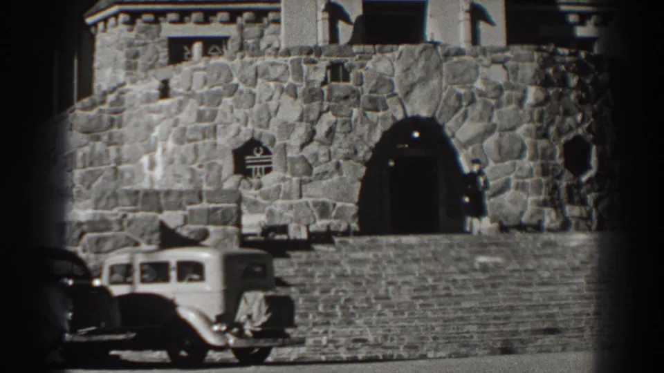 A stone facade at Bonneville Dam shows an arched entrance, rough masonry, a parked 1930s automobile, and a person near the doorway.