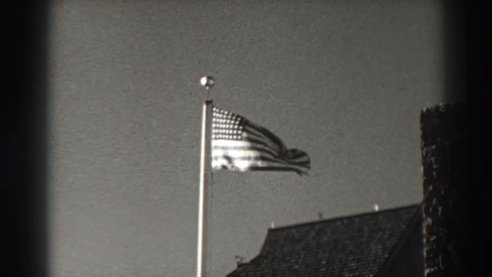 The flag stretches outward in the wind, framed by a roofline and dark foreground edge in Oregon archival footage.