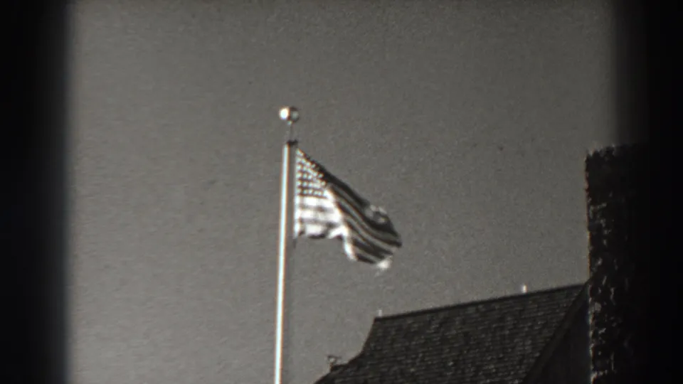 The U.S. flag folds back toward the pole above a roofline, with grain and soft monochrome contrast visible.