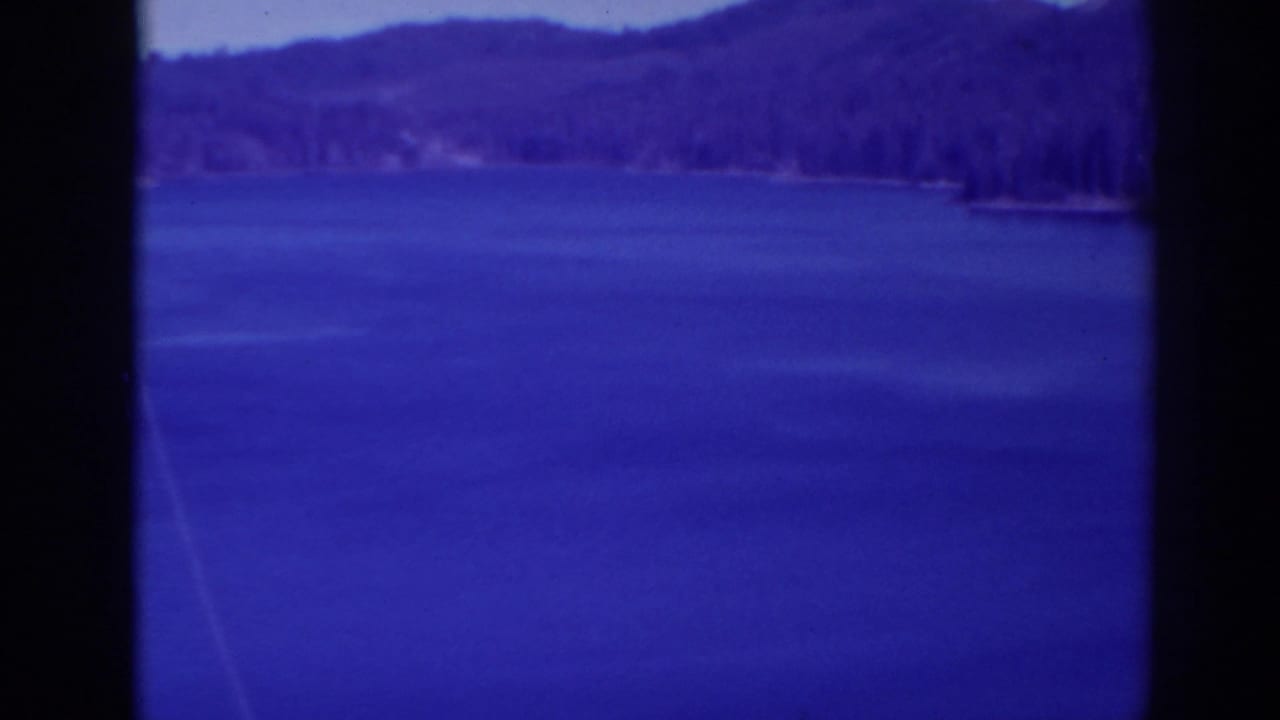 Blue lake water with forested mountains and pale sky beyond.
