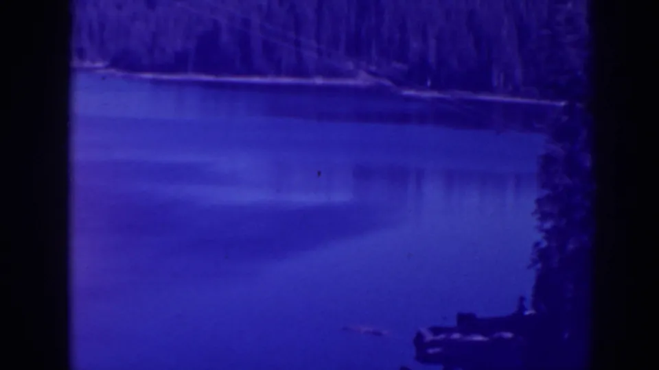 1938 home-movie view of Bucks Lake with calm water, evergreen shoreline, dark frame edge, and a faint object near the lower right.
