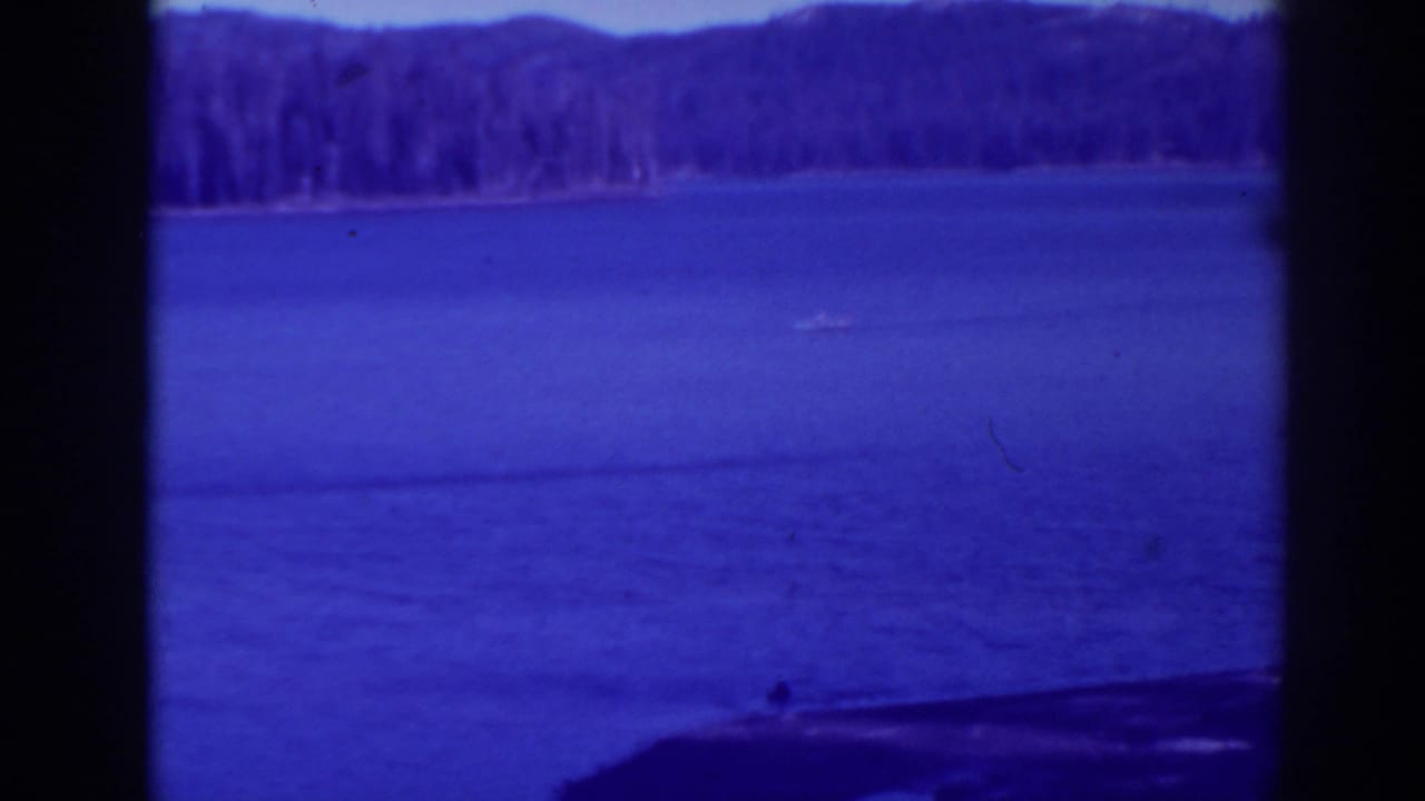 Small boat on blue lake water with distant forested shoreline and dark foreground framing.