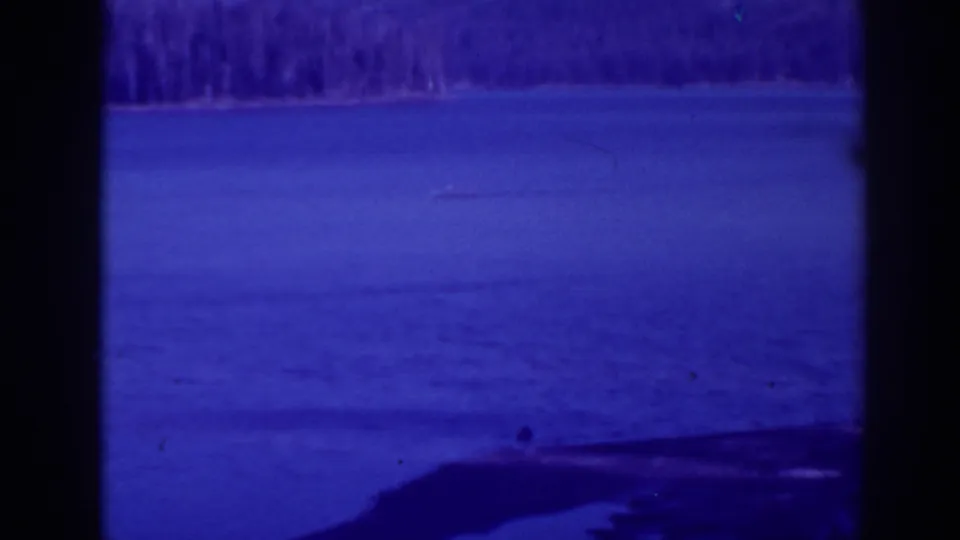 Blue-tinted 1938 home-movie frame of Bucks Lake with calm water, distant trees, and a dark shoreline or dam edge in the foreground.
