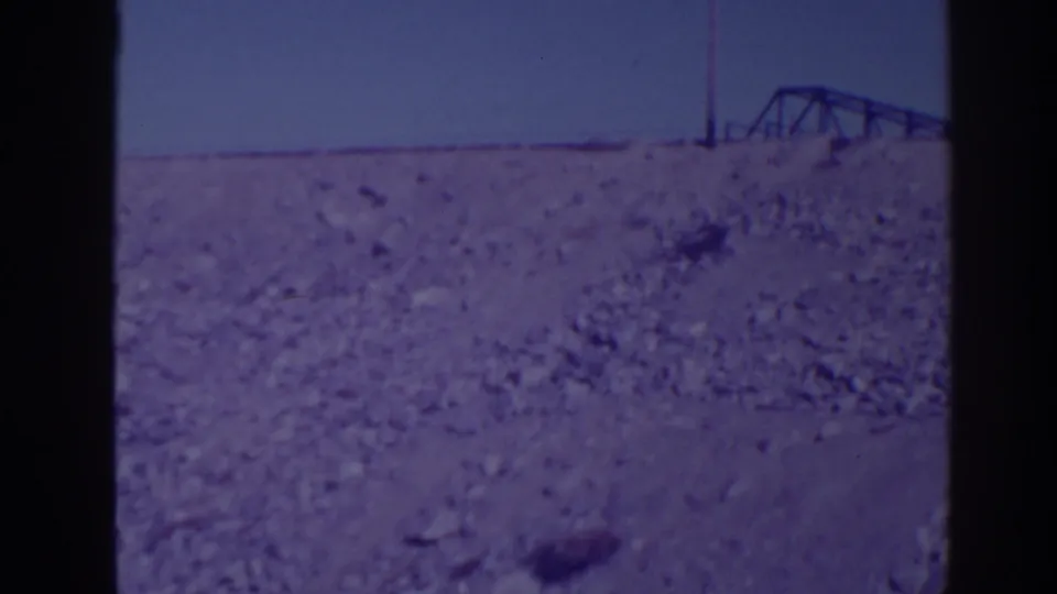 1938 Bucks Lake area frame showing barren rocky ground, a distant metal framework, and a utility pole against a pale sky.