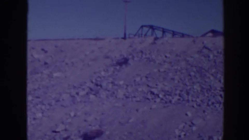 1938 Bucks Lake area frame with rocky foreground, distant utility pole, metal framework, and a small horizon structure in soft 8mm color.