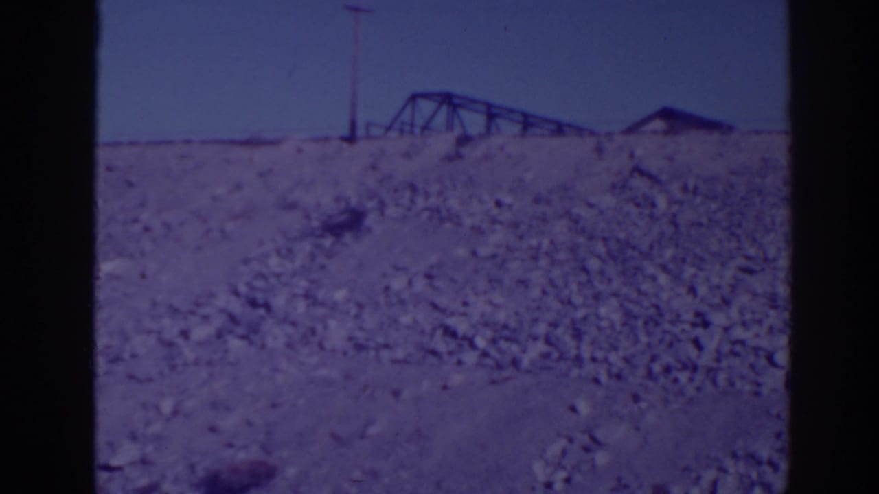 Rocky hillside with a distant truss structure, utility pole, small roofline, and clear sky.