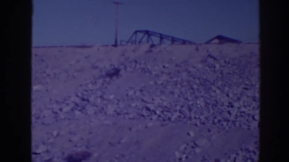 1938 Bucks Lake area view showing a rocky hillside, distant metal truss structure, utility pole, and small roofline under clear sky.