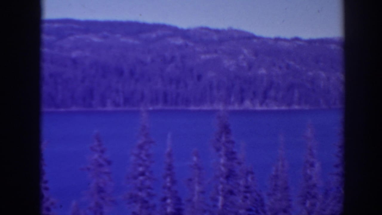 Blue lake with evergreen treetops and distant wooded hills, bordered by dark film edges.