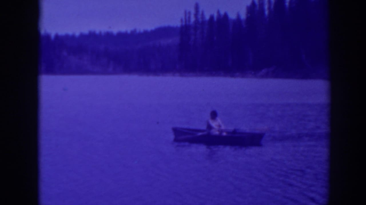 Lone person rowing a small boat across a calm lake near a dark forested shore.