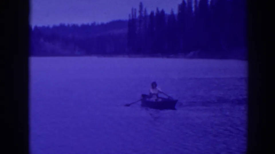 A distant adult rows a small boat on Bucks Lake, with soft 8mm grain, blue dusk tones, and a dark evergreen background.