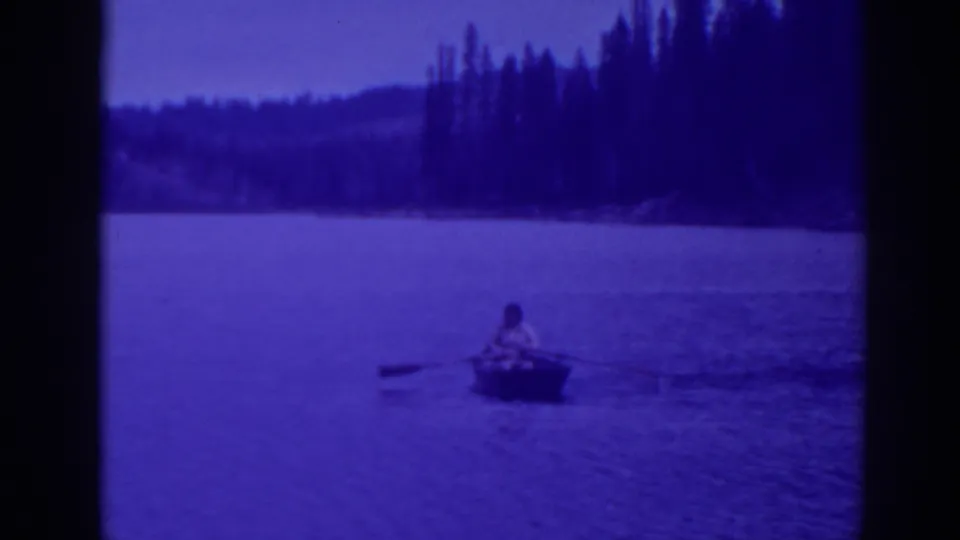 Bucks Lake 1938 frame with a lone adult rower centered on blue water below a dense evergreen shoreline.
