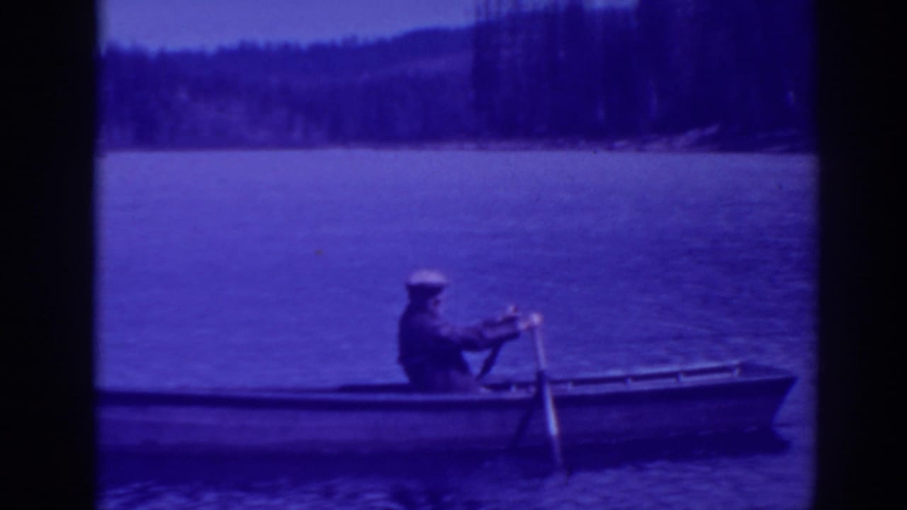 Man in a small rowboat on a calm lake with a forested shore behind him.