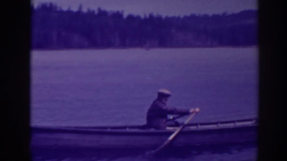 The rower passes through frame in a low wooden boat on Bucks Lake, with a dark tree line and aged purple 8mm color visible.