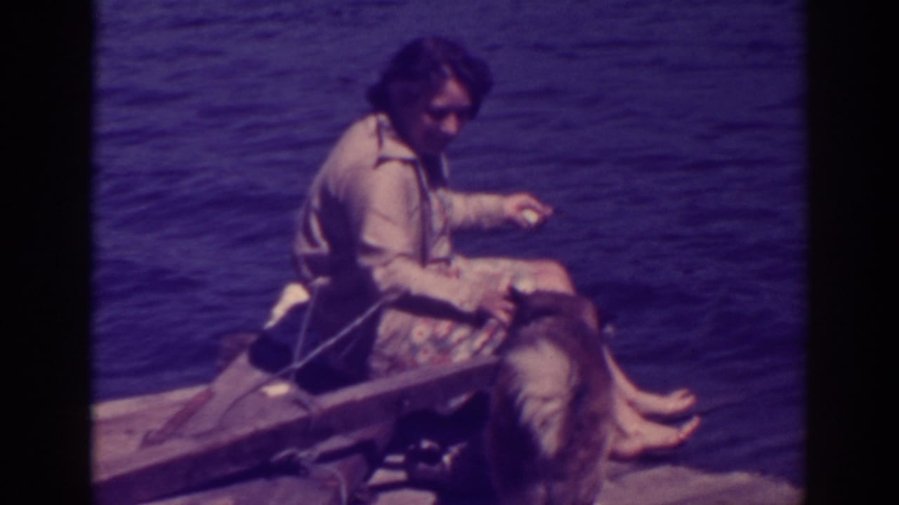 Woman seated barefoot on a wooden raft beside a large dog on lake water.