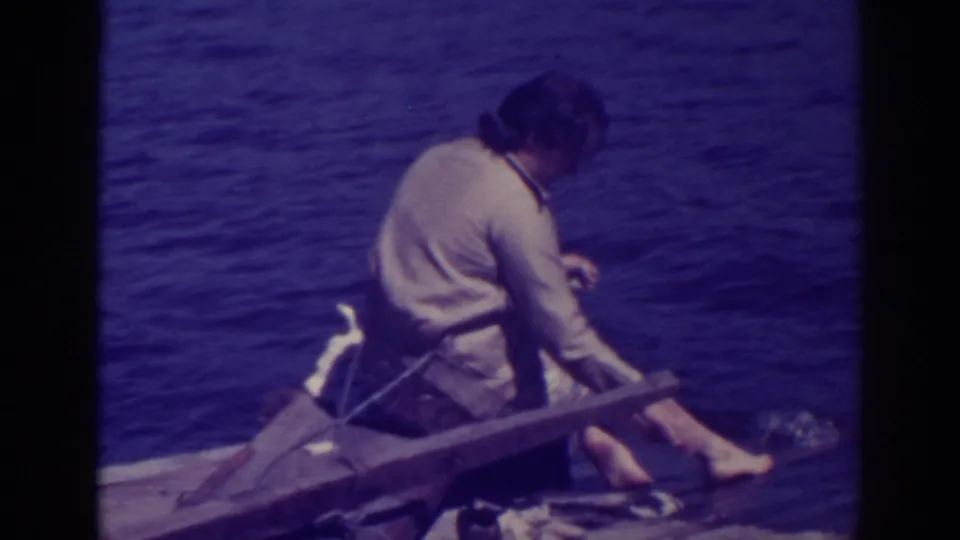 The 1938 home-movie frame shows the woman seated sideways on the dock, continuing to wash clothing at the lake edge.