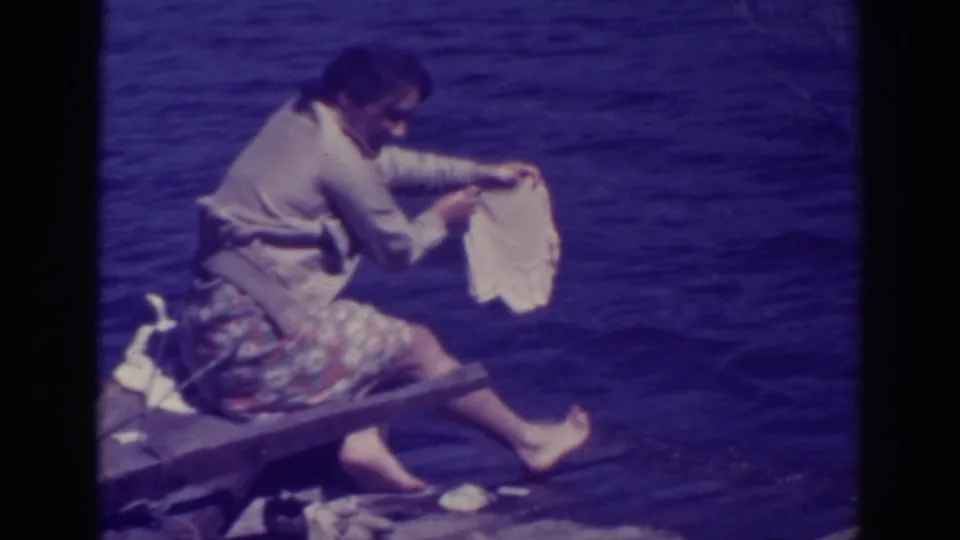At Bucks Lake, the woman lifts a pale garment from the water while seated barefoot on the wooden dock.