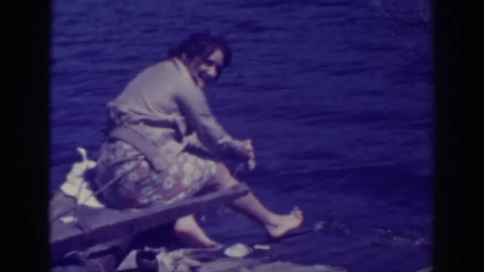 The woman glances back toward the camera as she rinses or wrings cloth at the Bucks Lake shore, with blue-purple aged 8mm color visible.