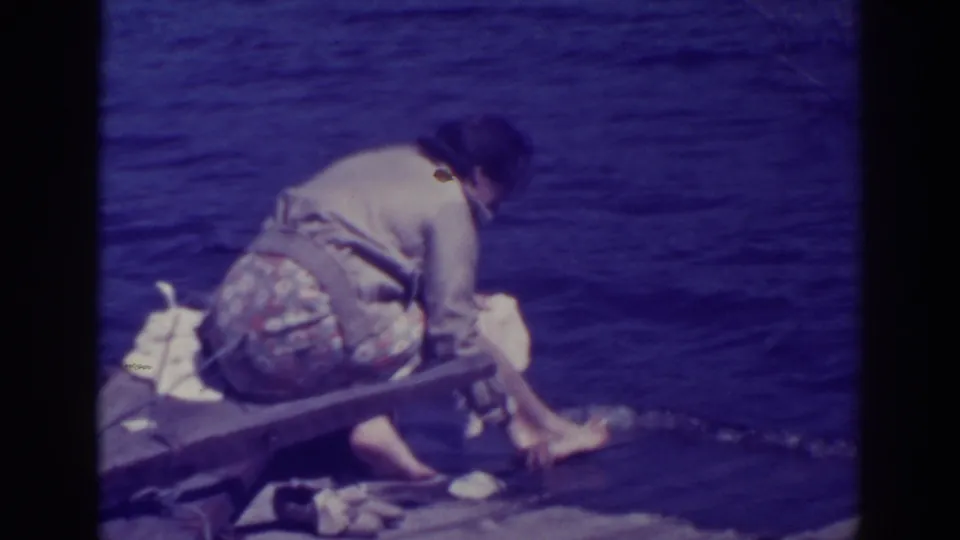 The woman leans forward from the bench or dock edge, lowering laundry toward the lake water along the rocky Bucks Lake shoreline.