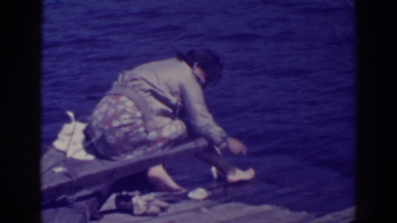 Woman crouching barefoot on a wooden pier beside dark blue lake water.