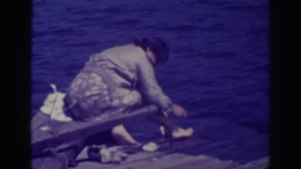 At Bucks Lake, California, in 1938, a woman in a floral skirt and apron crouches on a wooden pier while handling a fish at the water's edge.