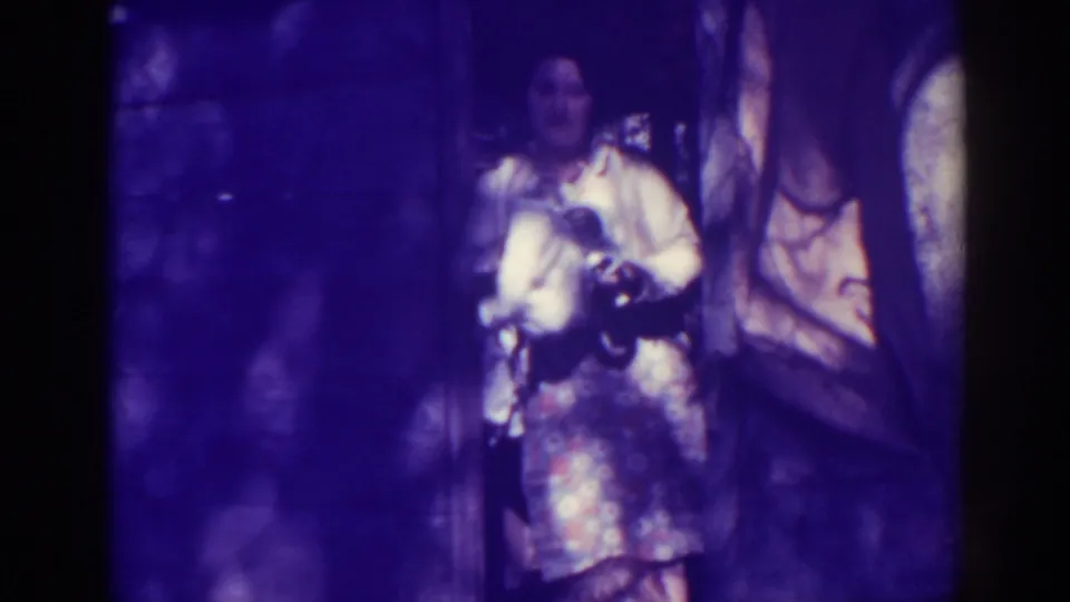 The woman remains in the rustic doorway, holding the kettle in front of her in purple-tinted 8mm home-movie footage.