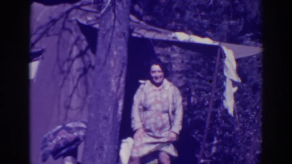 A woman in a patterned dress smiles toward the camera outside a tent at Bucks Lake, with laundry and trees around the campsite.