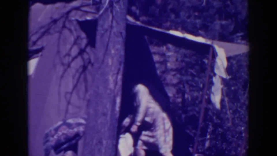 The woman bends down outside the makeshift tent at Bucks Lake, handling light-colored laundry beside the tree and shaded shelter.
