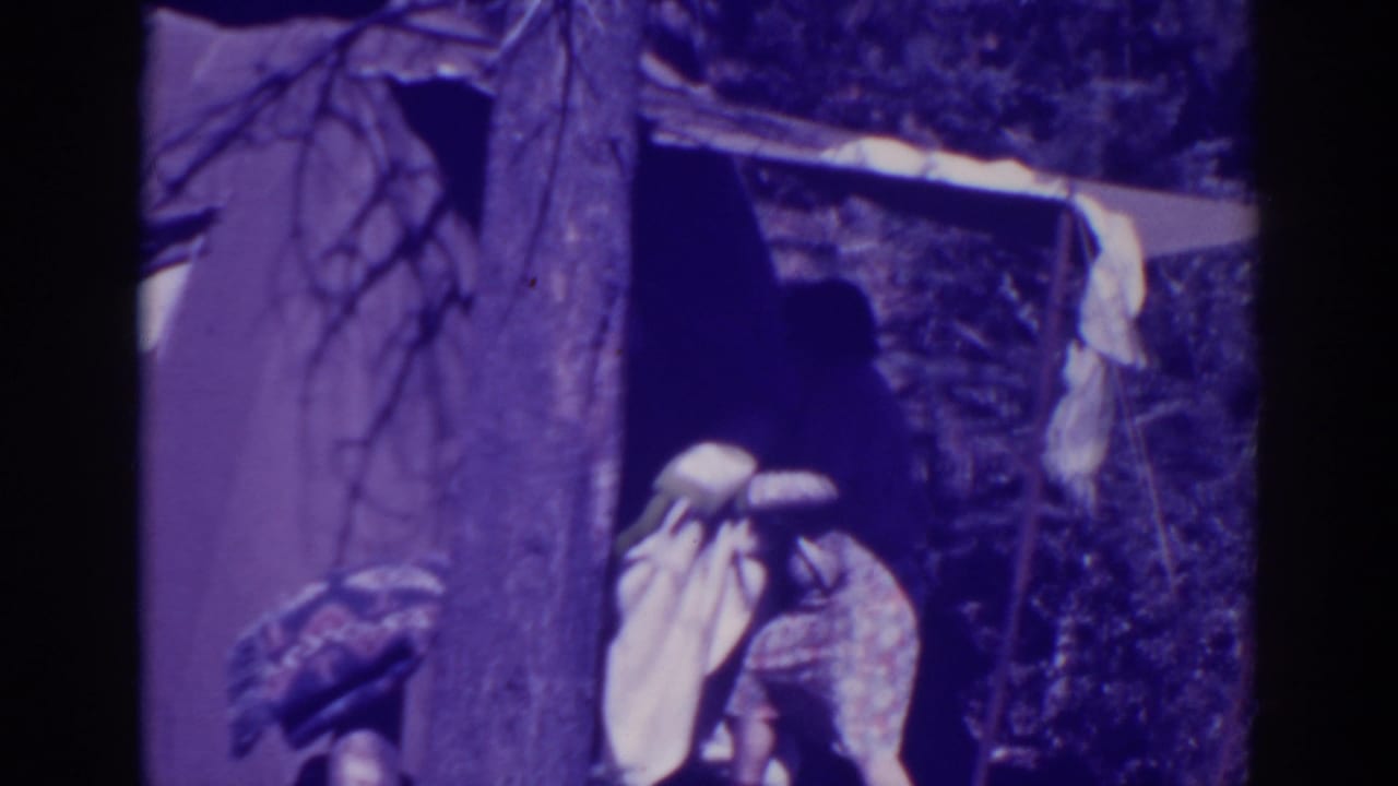 Person in light clothing handling a blanket beside a tent and tree trunk.