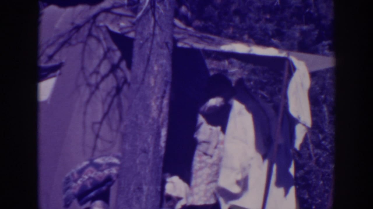 Woman in a light dress stands at a canvas tent beside a tree.