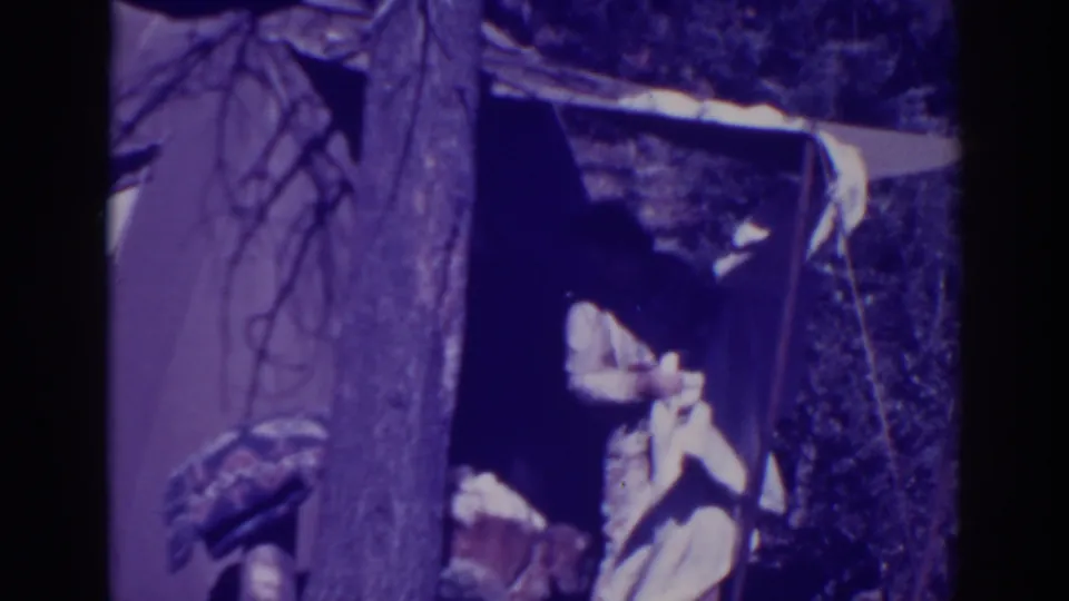The woman bends or leans near the tent opening while handling a light-colored item in the wooded campsite.