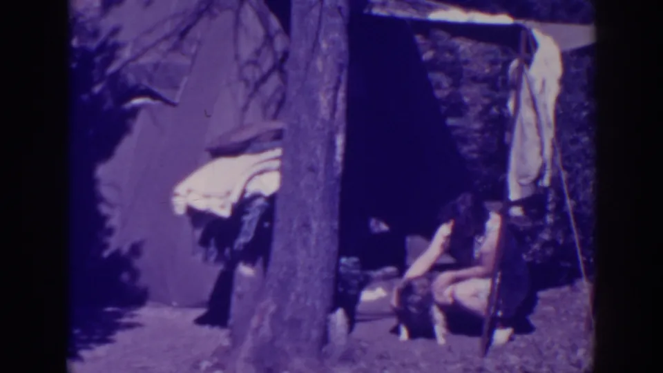 The 1938 home-movie frame shows the woman bending toward the dog at a Bucks Lake campsite with shelter fabric and blankets nearby.