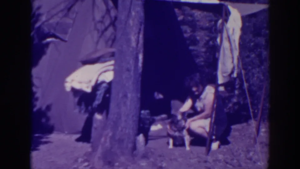 A dog stands beside the crouching woman at the Bucks Lake campsite, framed by a tree, blankets, and a dark shelter wall.