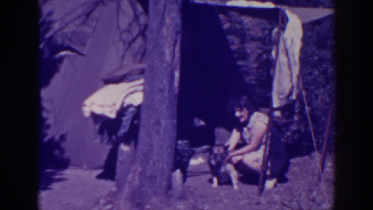 Woman crouches beside a small dog near a canvas tent and large tree trunk.