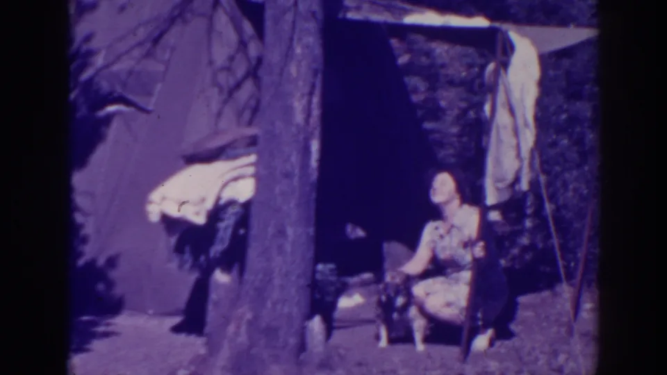 At the campsite, the woman looks upward while crouched beside the small dog, with the tent wall and tree trunk dominating the frame.