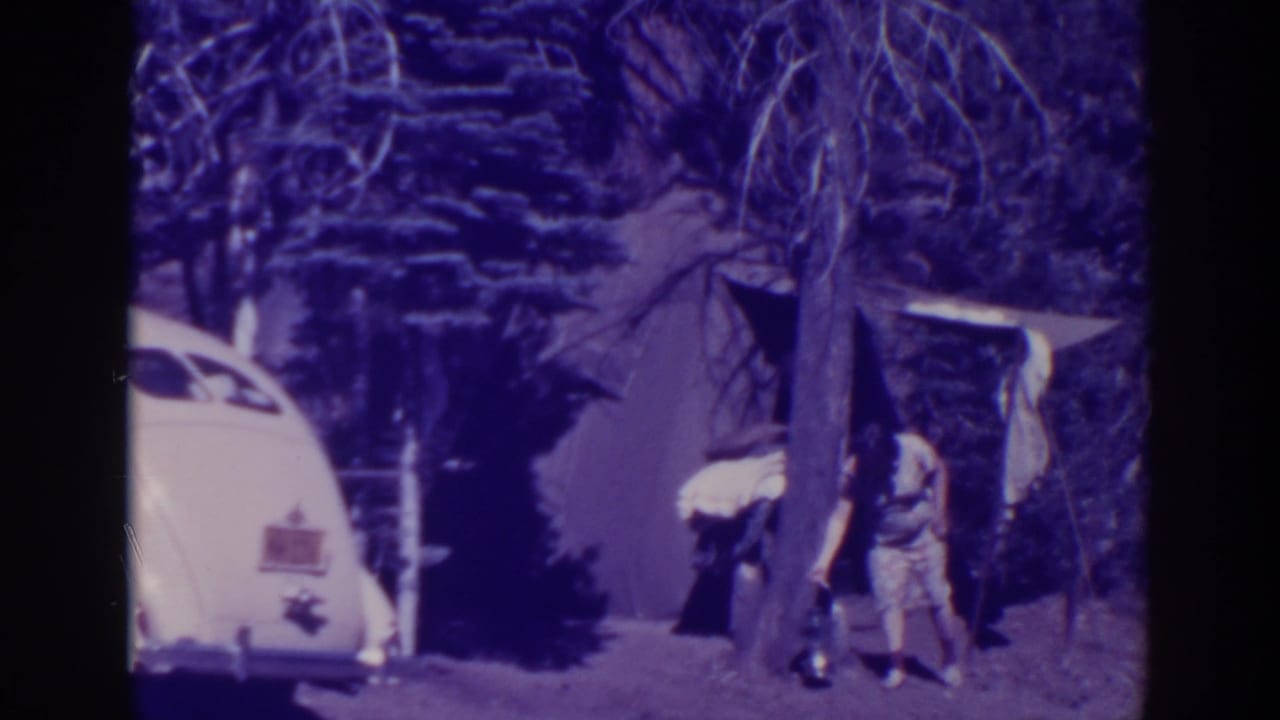 Adult bending near a canvas tent with a white car and trees visible at the campsite.