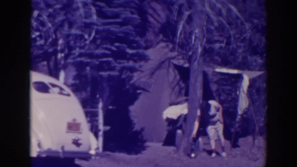 A person bends near a tree and canvas tent at the Bucks Lake forest campsite, with the rounded white car partly visible.