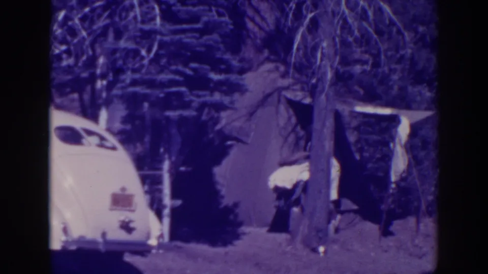 The campsite view holds on the tent and white car as the woman moves out of clear view among trees and tent fabric.