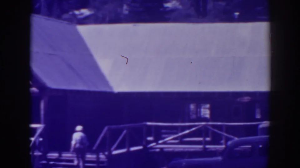 Purple-blue 8mm frame of the Bucks Lake wooden structure with roofline, porch rail, steps, a distant person, and part of a parked car.